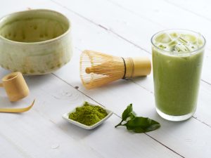 Iced Matcha Latte Tea and homemade tea brewing accessories with bamboo whisk, green tea powder on white wooden background.